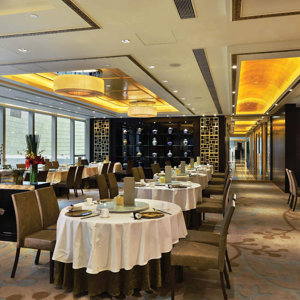 Aedas Interiors’ Ming Court at Langham Place, Hong Kong is shown as an upscale Chinese fine‑dining interior with round banquet tables, upholstered chairs, and a refined palette of warm neutrals. The image highlights silver‑leaf accented wall panels, abstract floor patterns inspired by Chinese characters, illuminated ceiling coffers, and a carefully balanced blend of traditional motifs with contemporary luxury finishes.