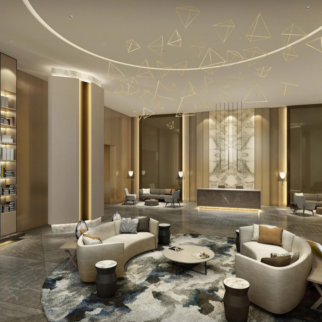 Aedas Interiors’ MIG Colony Residential Project in Mumbai is shown as an elegant residential lobby with curved upholstered seating, a sculptural feature chandelier composed of delicate geometric forms, and warm timber‑lined walls. The image highlights a contemporary living environment defined by soft neutral tones, layered lighting, polished stone flooring, and refined lounge configurations that create a calm, aspirational communal arrival space within a high‑density residential development.