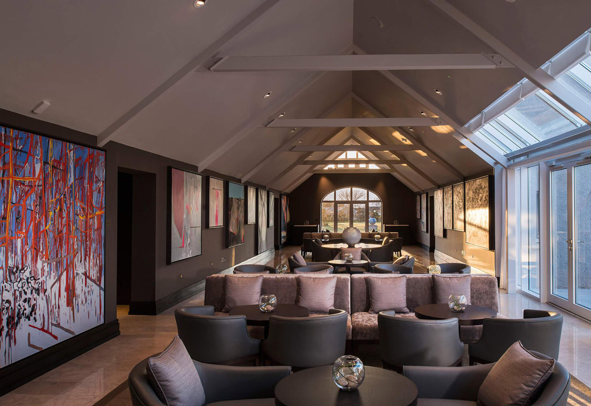 Aedas Interiors’ Twr y Felin Hotel in St Davids is shown as an elegant gallery lounge with pitched timber roof trusses, skylight glazing, polished stone flooring, and curated contemporary artworks lining dark feature walls. The image highlights a refined hospitality interior that merges historic architectural fabric with modern bespoke furnishings, creating an intimate, art‑driven sanctuary with strong visual focus and layered seating zones.
