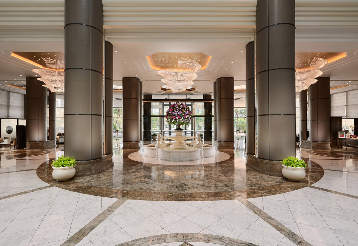The renovated Grand Hyatt Taipei lobby is shown as a grand, symmetrical arrival hall with tall cylindrical columns, polished marble flooring, and layered coffered ceilings. Large sculptural crystal chandeliers hang above a central floral arrangement, while warm lighting and refined stone finishes create a luxurious, contemporary hospitality interior with a strong sense of scale and ceremonial arrival.