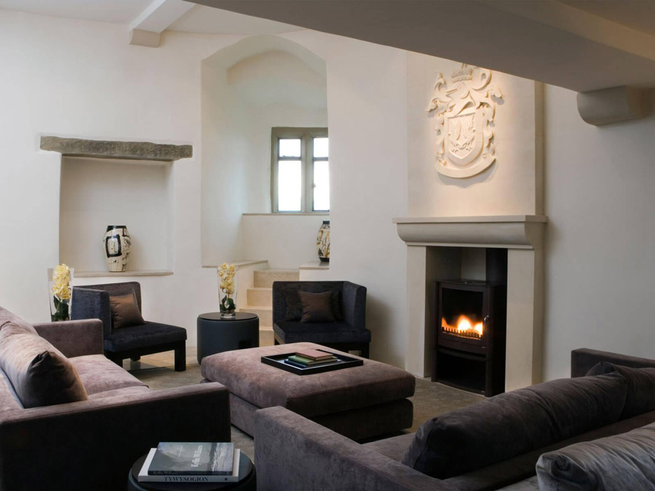 Aedas Interiors’ Roch Castle Hotel in Haverfordwest is shown as an intimate historic lounge with thick whitewashed stone walls, a sculpted stone fireplace, soft upholstered seating, and a glowing hearth. The image highlights a carefully restored medieval interior where restrained contemporary furnishings, heritage masonry, and curated artifacts create a calm, atmospheric hospitality space that foregrounds the castle’s history and sense of sanctuary.