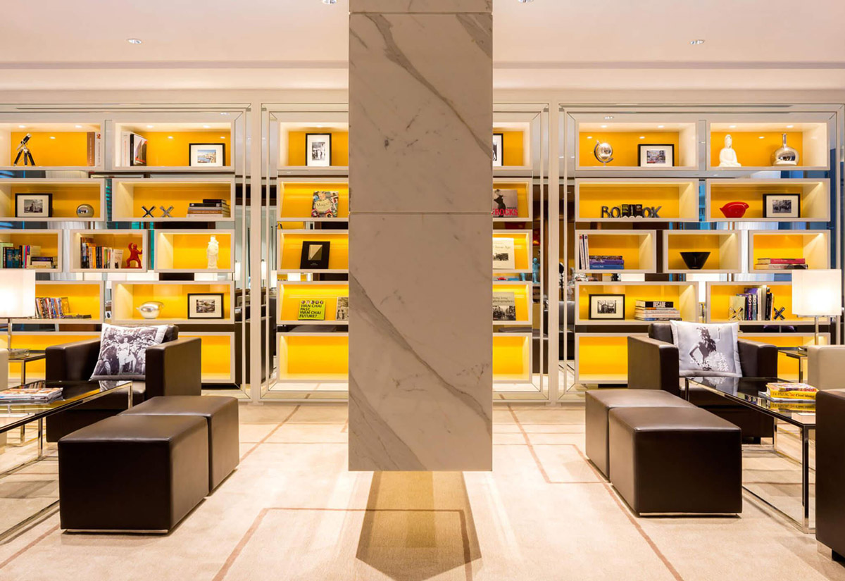 Aedas Interiors’ Hotel Indigo Hong Kong Island is shown as an intimate lounge setting with symmetrical built‑in shelving illuminated in warm yellow, marble column framing, and low leather seating. The image highlights a boutique hospitality interior that layers curated objects, graphic books, and soft lighting to express local character while maintaining a refined, residential‑style atmosphere within a dense urban hotel environment.