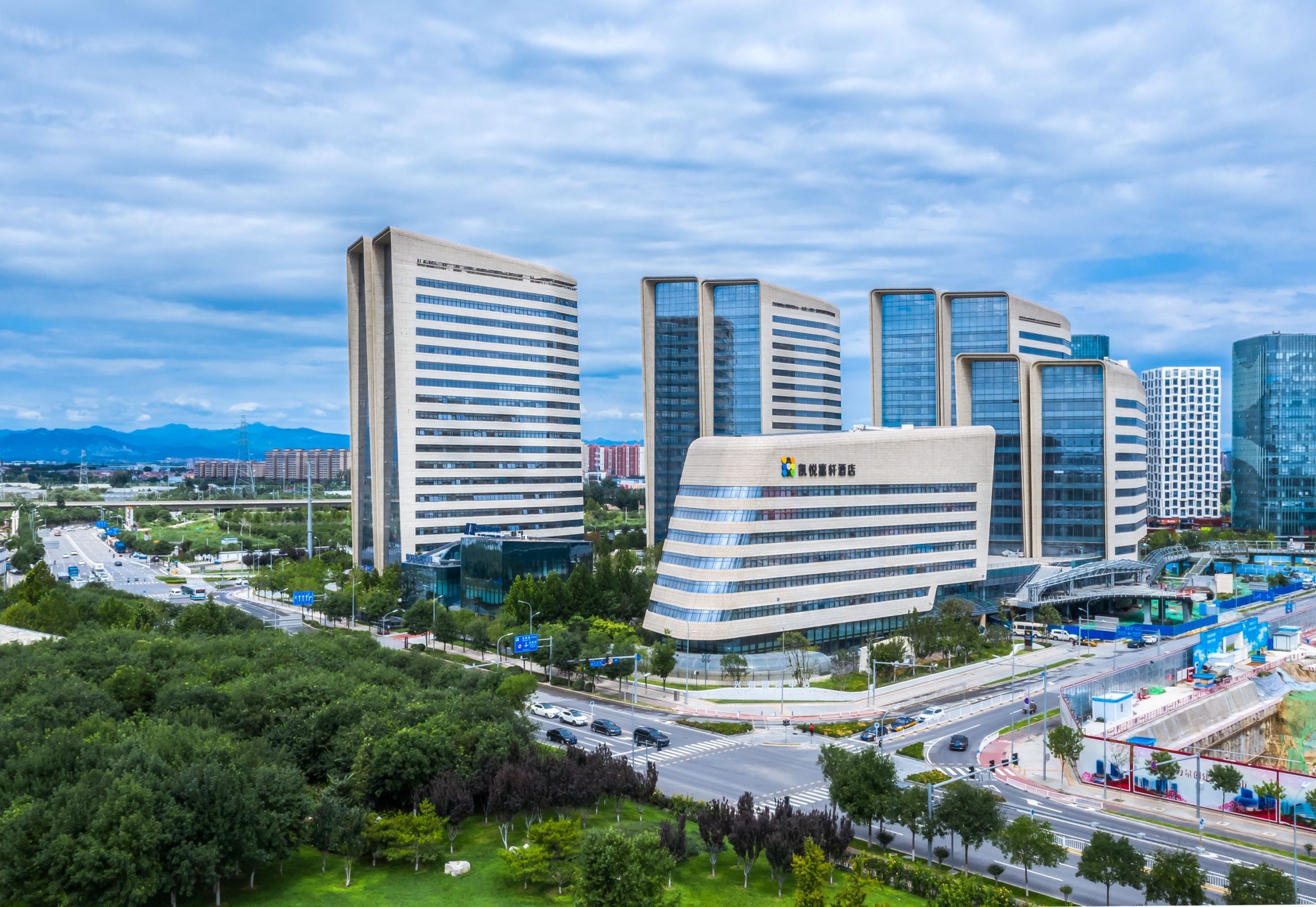 Aedas’s Hyatt Place Hotel at Daxing Xihongmen is shown as a large mixed‑use development with vertically oriented office towers and a horizontally layered podium accommodating retail and hotel functions. The aerial view highlights striated façade articulation, a strong urban edge to the north, and integration with adjacent green space.