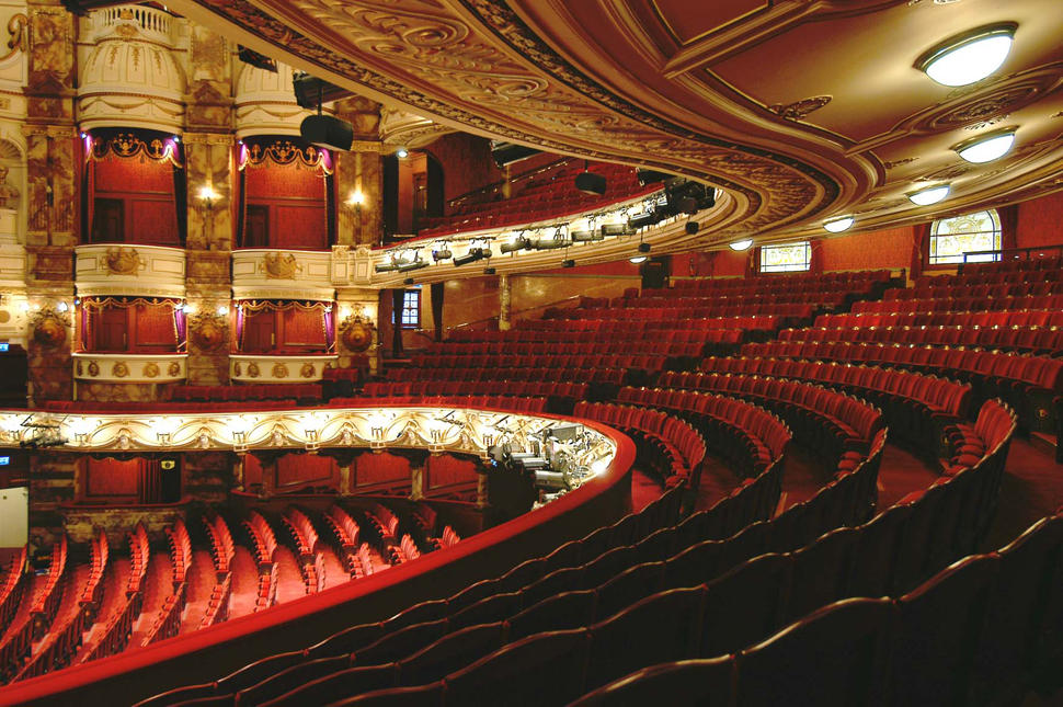 Aedas’s London Coliseum is shown as a large historic theatre architecture with a sweeping auditorium section, curved balcony tiers, ornamental structural envelopes, and a deep proscenium opening. The image highlights heritage performance architecture, large‑span spatial volume, and building‑scale acoustic enclosure within a West End cultural landmark.