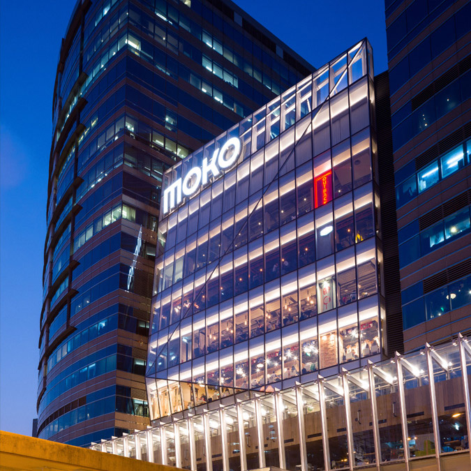 Aedas’s MOKO in Hong Kong is shown as a mixed‑use commercial building with a transparent glazed retail podium, illuminated multi‑storey façade, and an attached curvilinear office or hotel tower. The night‑time exterior highlights curtain‑wall architecture, vertical stacking, and integration within a dense urban context.