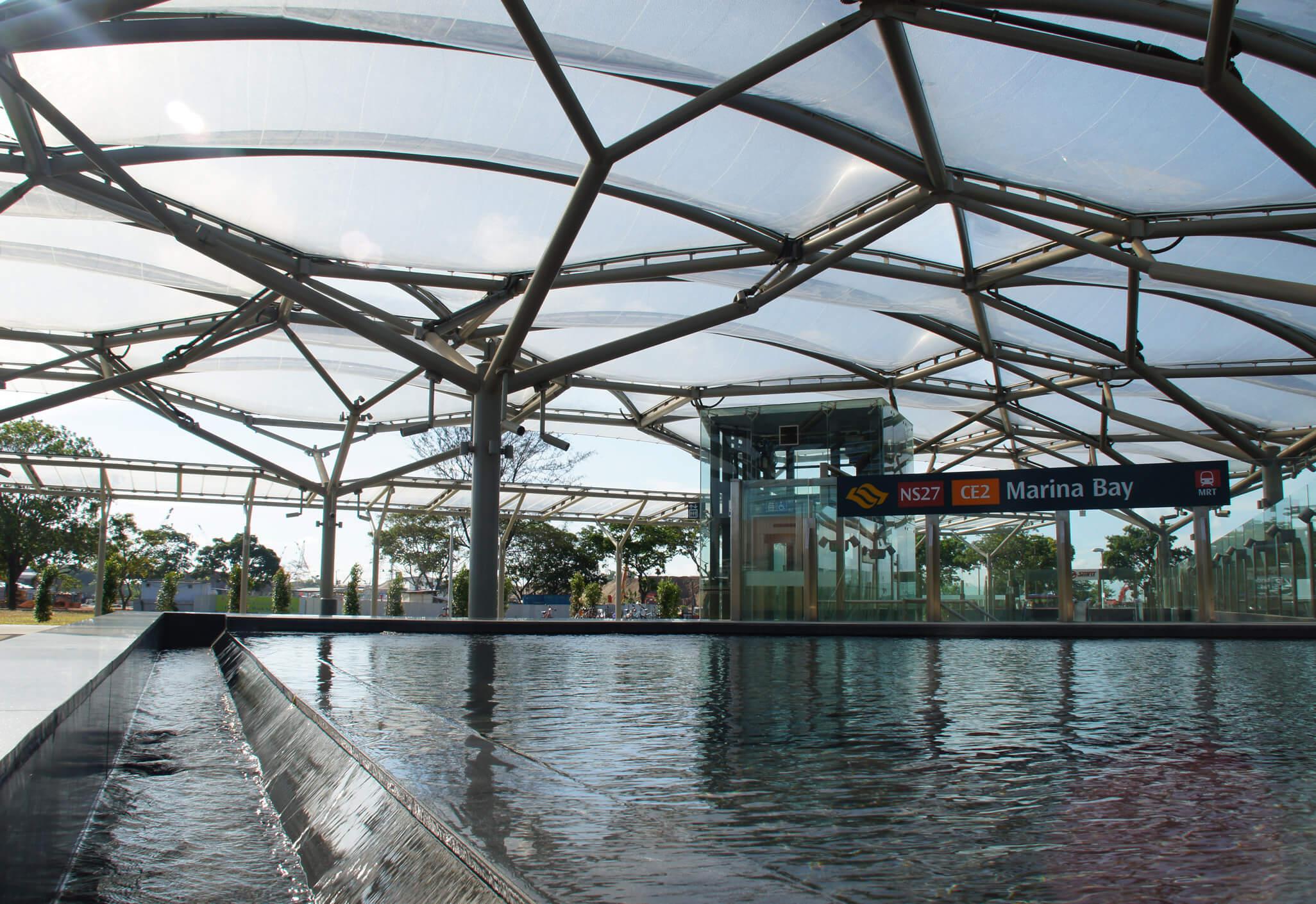 Aedas’ Marina Bay MRT Station with ETFE skylight canopy, reflective pool and glazed entrance set within parkland. Street‑level view of the Aedas‑designed Marina Bay MRT Station in Singapore, showing a lightweight steel structure supporting a translucent ETFE canopy. The canopy spans above a reflective pool and glass lift enclosure, allowing daylight penetration while maintaining visual openness to the surrounding park landscape in a tropical urban environment.