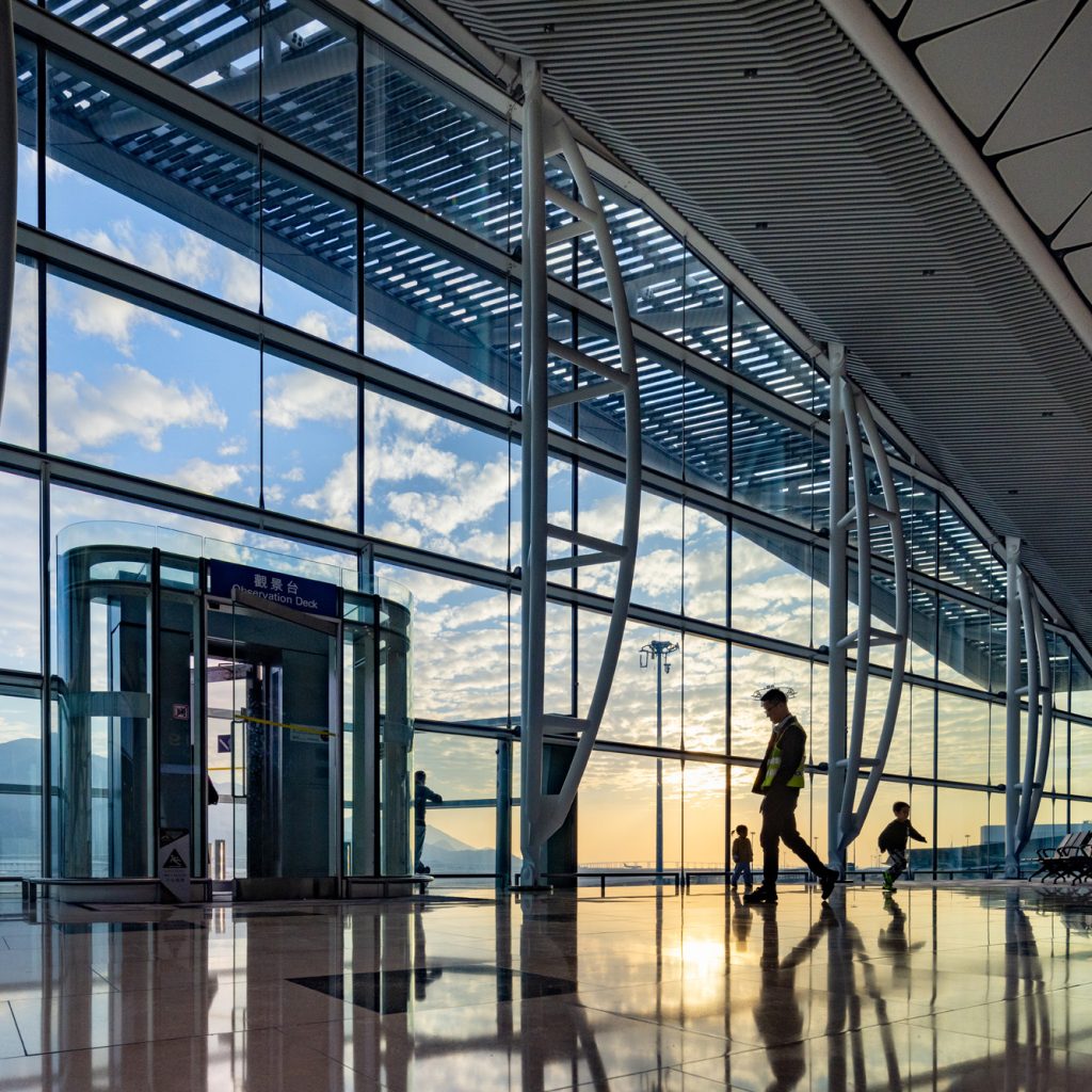 Aedas’ Midfield Concourse at Hong Kong International Airport with full‑height glazed façade, steel mega‑columns and rooflights. Interior view of Aedas-designed Midfield Concourse at Hong Kong International Airport, showing a full-height glazed façade with external shading, inclined steel mega-columns, polished stone flooring and integrated rooflights. Daylight penetrates deep into the five-level concourse, supporting clear passenger circulation, automated people mover access and long-span terminal operations within a subtropical climate.
