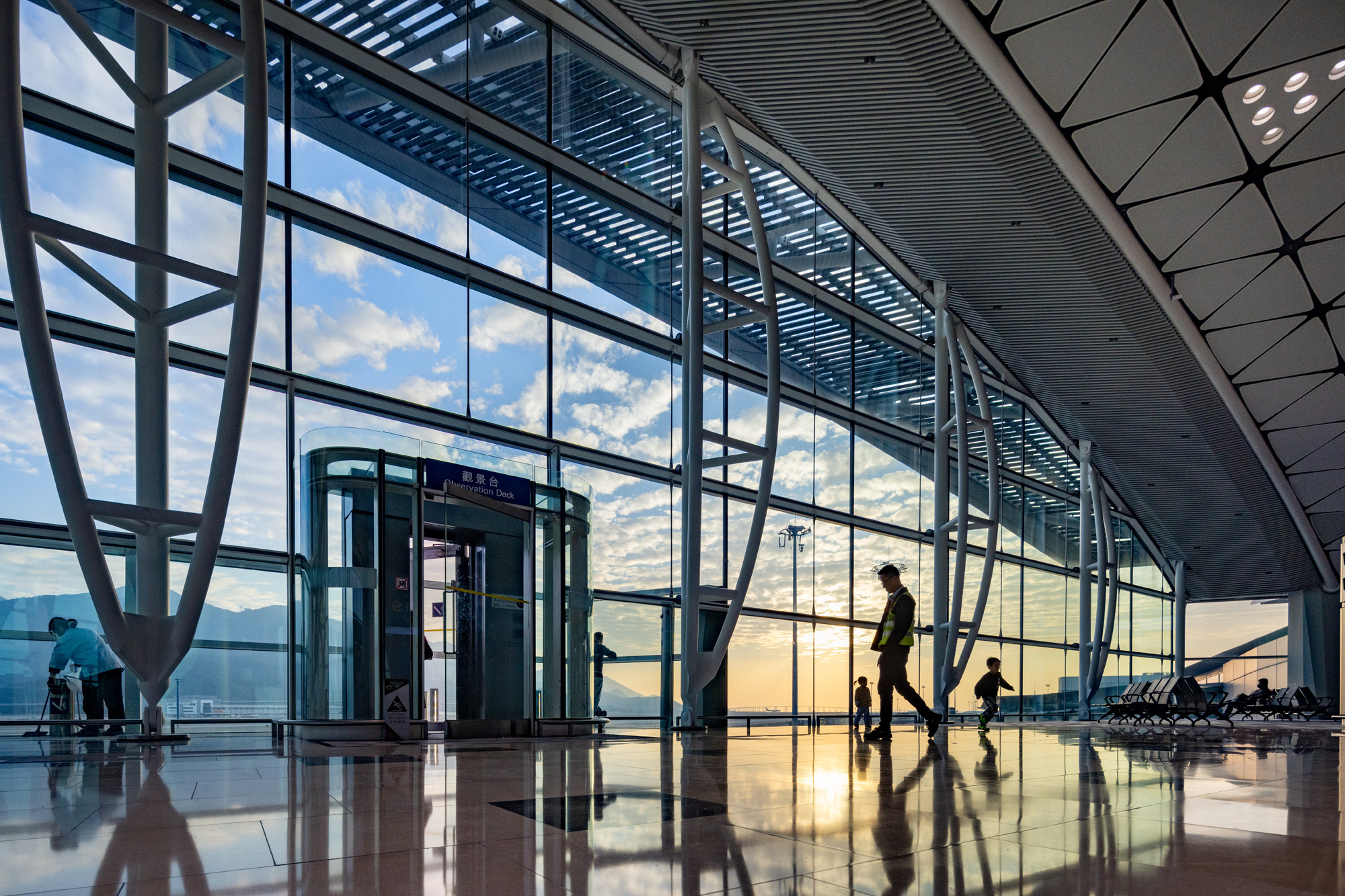 Aedas’ Midfield Concourse at Hong Kong International Airport with full‑height glazed façade, steel mega‑columns and rooflights. Interior view of Aedas-designed Midfield Concourse at Hong Kong International Airport, showing a full-height glazed façade with external shading, inclined steel mega-columns, polished stone flooring and integrated rooflights. Daylight penetrates deep into the five-level concourse, supporting clear passenger circulation, automated people mover access and long-span terminal operations within a subtropical climate.