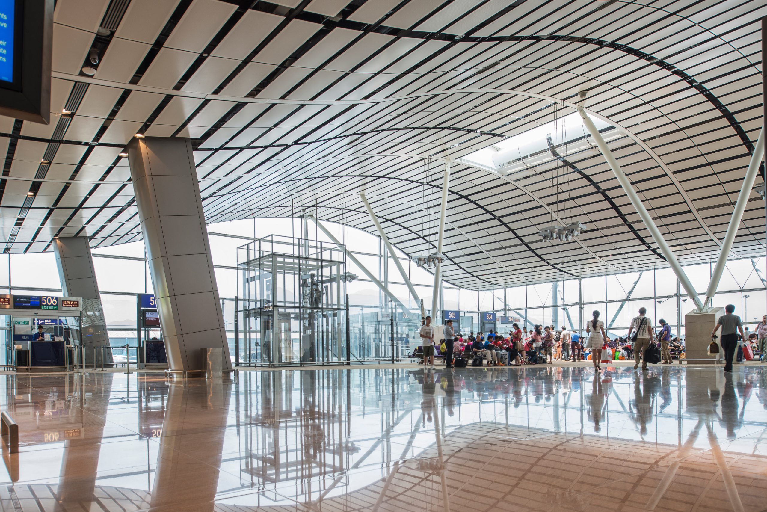 Aedas’ North Satellite Concourse at Hong Kong International Airport, showing winged roof, exposed steel columns and full‑height glazing. Interior view of Aedas-designed North Satellite Concourse at Hong Kong International Airport, featuring a curved winged roof with linear ceiling baffles, inclined steel support columns and full-height glazing. Polished stone flooring reflects natural daylight across the open passenger hall, with seating zones, vertical circulation cores and clear sightlines supporting transfer and boarding operations.