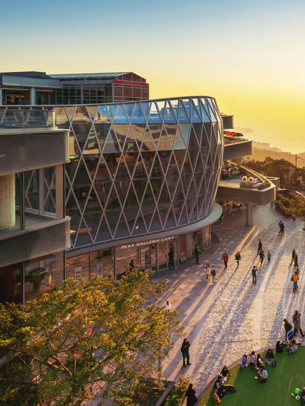 Aedas’s Peak Galleria in Hong Kong is shown as a hilltop retail destination with a faceted glazed façade, elevated pedestrian plaza, and stepped circulation. The image highlights transparent enclosure systems, public‑realm integration, and landscape‑responsive architecture positioned along Victoria Peak’s transport and viewing corridor.