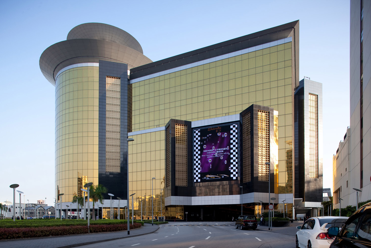 Aedas’ Sands® Macao casino resort with gold‑toned glazed façades, cylindrical hotel tower and podium entry. Street‑level view of the Aedas‑designed Sands® Macao showing a Las Vegas‑style casino podium clad in gold‑tinted curtain wall glazing, articulated by vertical black stone elements and a cylindrical tower volume above. A recessed vehicular drop‑off, landscaped forecourt and broad roadway frame the building within Macau’s dense urban resort context.