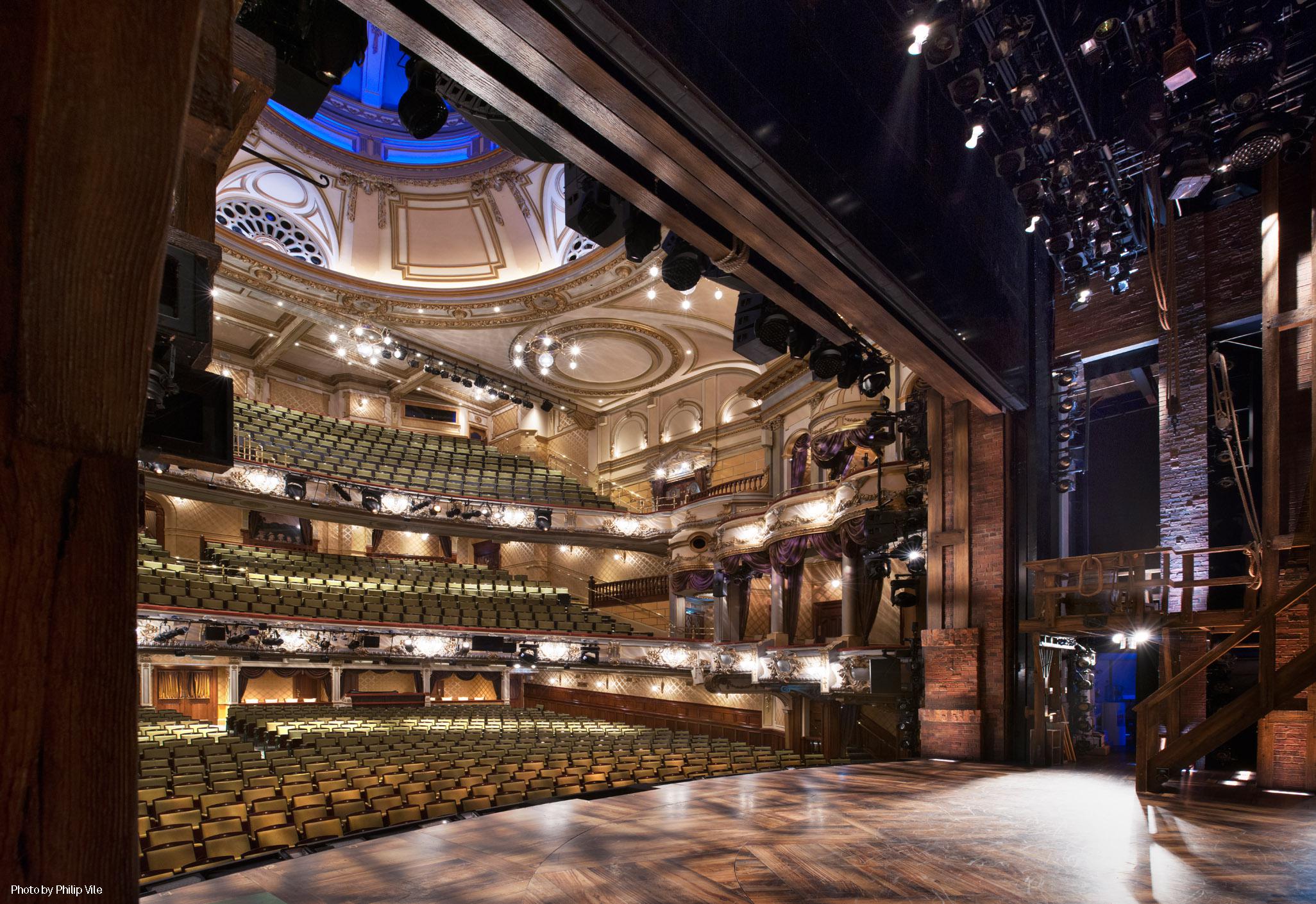 Aedas’s Victoria Palace Theatre in London is shown as a historic proscenium theatre architecture with a deep stage house, multi‑tiered balconies, ornamental structural detailing, and integrated lighting infrastructure. The image highlights heritage theatre massing, sectional organization, and performance‑led architectural volume within a dense West End context.