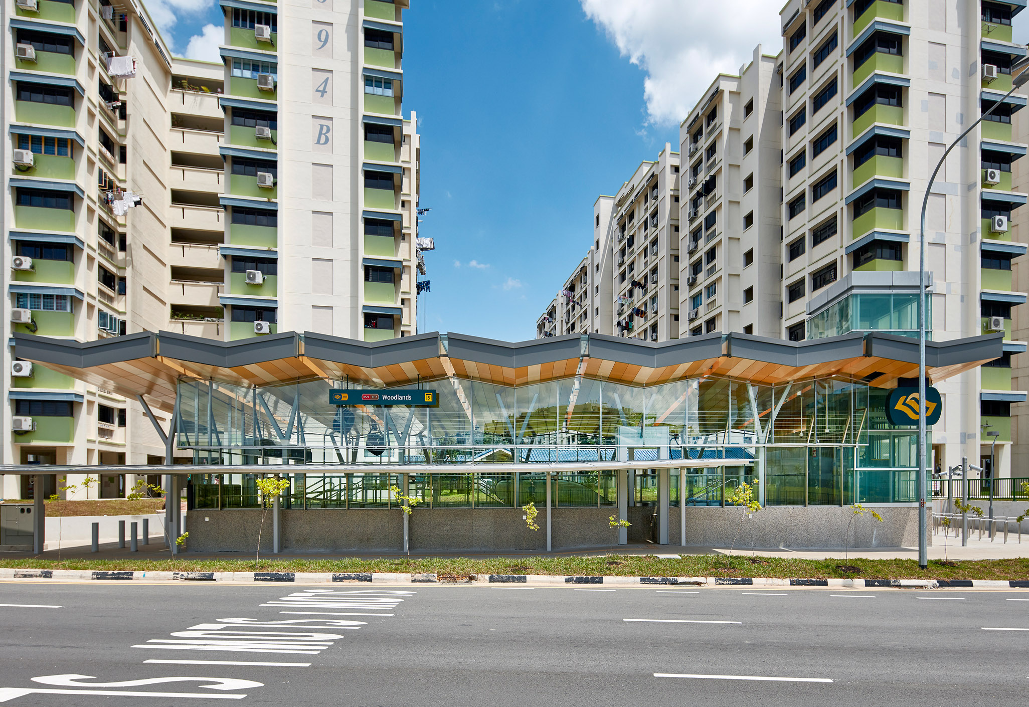 Aedas’s Woodlands Station on Singapore’s Thomson–East Coast Line is shown with a transparent glazed station box, slender steel supports, and a folded lightweight roof canopy. The exterior highlights climate‑responsive transit architecture, clear structural expression, and integration with surrounding high‑density residential buildings in a tropical urban context.
