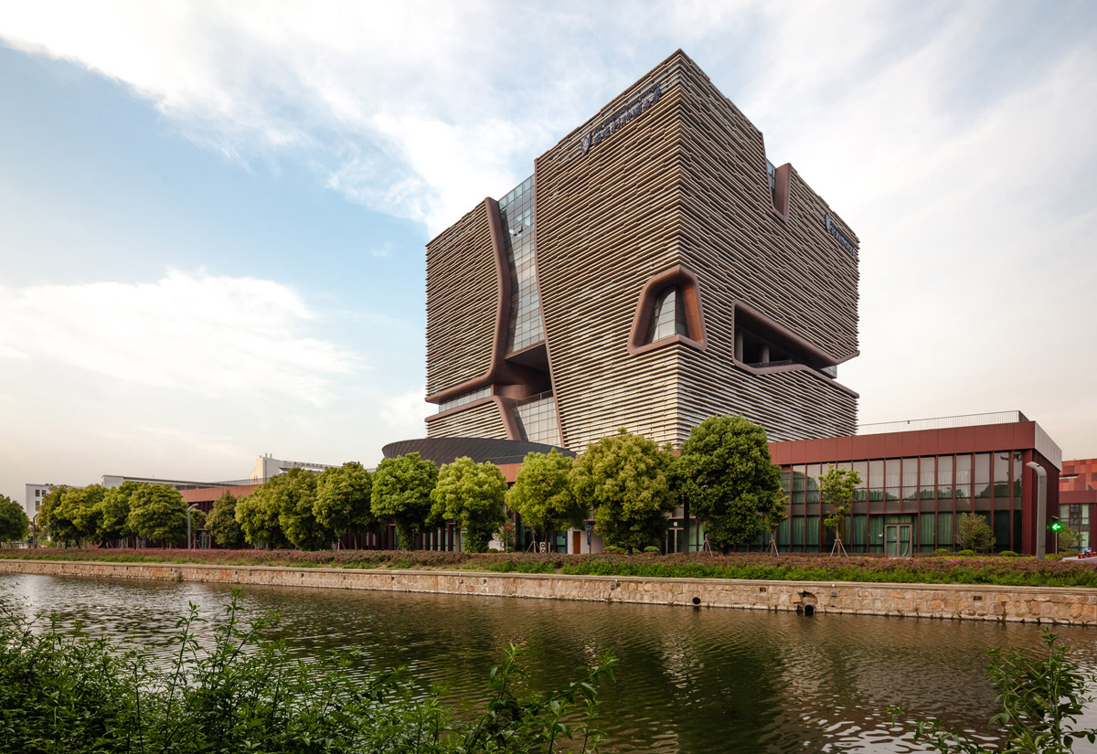 Aedas’ Jiaotong-Liverpool University Central Building in Suzhou with porous cube massing, horizontal cladding and wind‑ventilating façade voids. External view of Aedas-designed Xi’an Jiaotong‑Liverpool University Central Building in Suzhou, showing a horizontally stratified cubic volume on a landscaped plinth. The façade is wrapped in layered horizontal fins, punctured by deep sculpted voids and slots that introduce daylight and enable passive airflow through the academic building in a temperate East China climate.