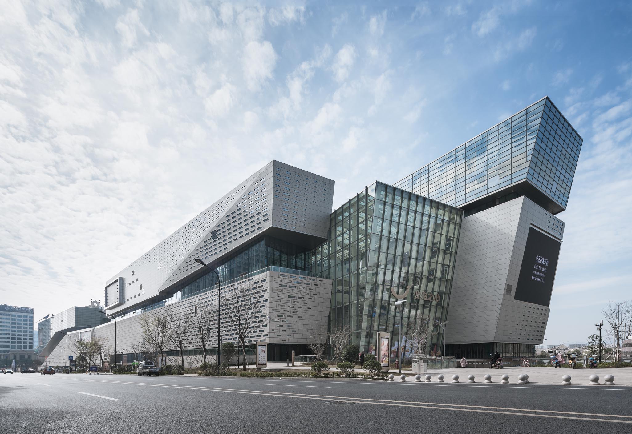 Aedas’s The Heart of Yiwu is shown as a large mixed‑use retail complex with bold angular building volumes, perforated metal façade panels, and a multi‑storey glazed atrium linking the primary blocks. The exterior highlights sculpted massing, structural clarity, and integration with a major urban roadway in Yiwu.