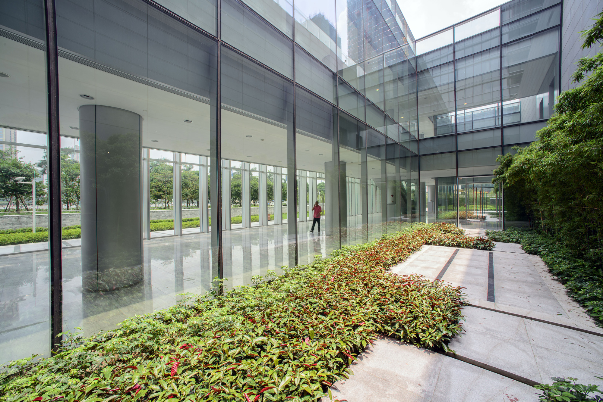 Aedas’ One AIA Financial Center in Foshan is shown with a glazed internal courtyard and circulation walkway featuring floor‑to‑ceiling glass façades, polished stone flooring, and integrated landscape planting beds. The image highlights a daylight‑filled transitional space where greenery, transparency, and reflective surfaces enhance spatial permeability, break down podium massing, and create a calm, biophilic environment for workplace interaction and movement.