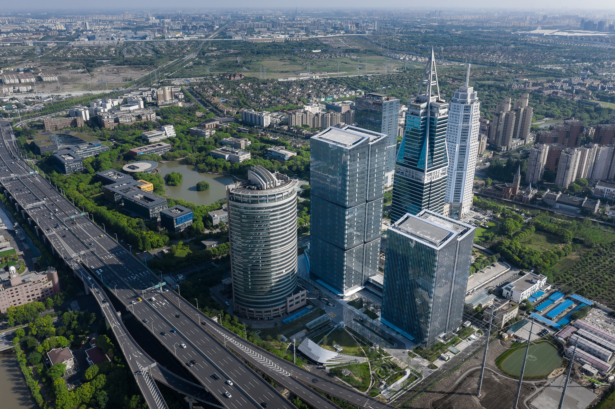 Aedas’ Shanghai International Fortune Centre is shown as a twin‑tower office development linked by a three‑storey podium, set within a dense green urban context. The aerial view highlights two glazed high‑rise towers with clean rectilinear forms, a bridging podium accommodating retail and F&B uses, and landscaped open spaces that integrate the complex with surrounding transport infrastructure and waterways in Jinqiao, Pudong.