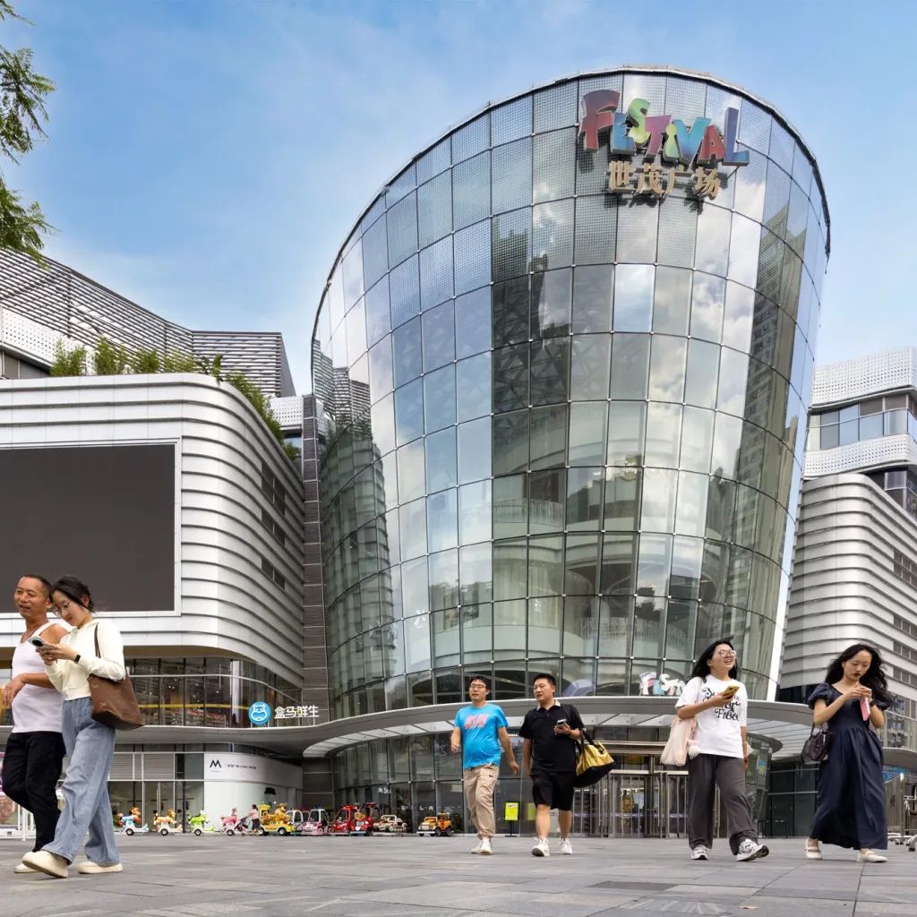 Aedas’s Chengdu Shimao Festival City is shown as a large mixed‑use commercial complex with a prominent cylindrical glass atrium, horizontally layered retail volumes, and curvilinear façade bands. The night‑time exterior highlights transparent curtain walls, multi‑level retail massing, and integration with a public urban plaza in Chengdu.