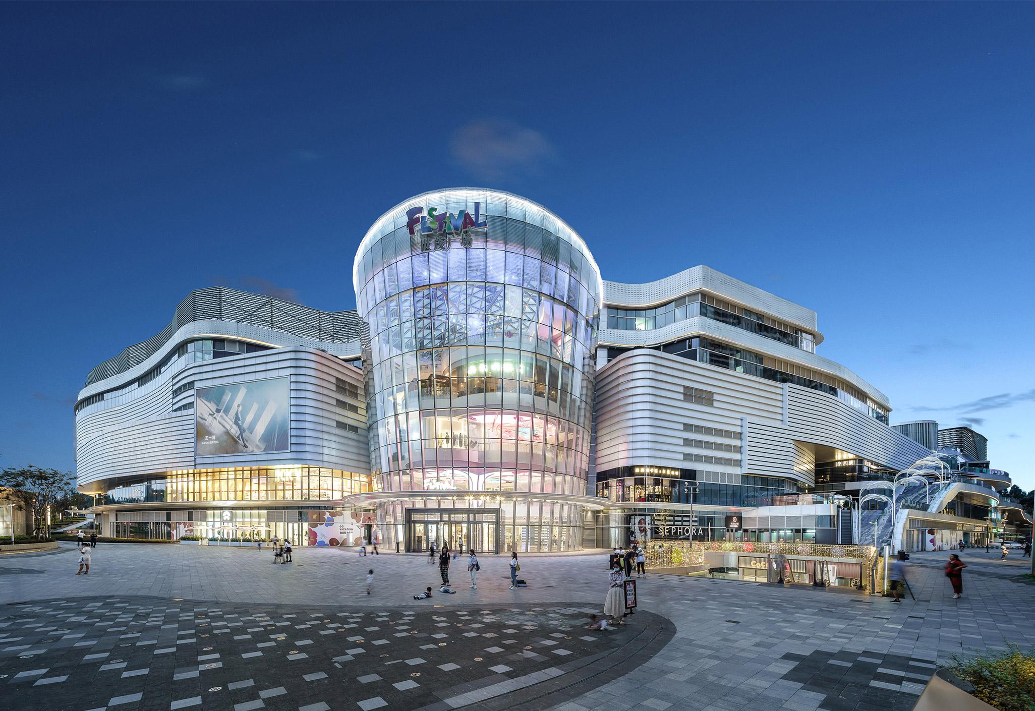 Aedas’s Chengdu Shimao Festival City is shown as a large mixed‑use commercial complex with a prominent cylindrical glass atrium, horizontally layered retail volumes, and curvilinear façade bands. The night‑time exterior highlights transparent curtain walls, multi‑level retail massing, and integration with a public urban plaza in Chengdu.