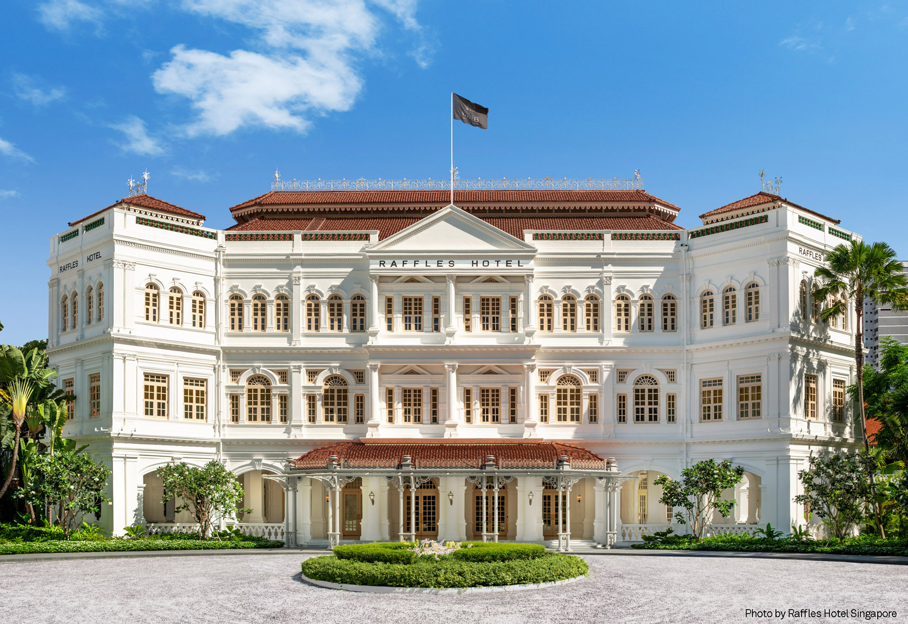 Aedas’ restoration of Raffles Hotel Singapore is shown as the iconic neoclassical façade with white colonnades, arched windows, and red‑tiled roofs set within lush tropical landscaping. The image highlights the carefully preserved heritage architecture, symmetrical proportions, and refined detailing that reflect the hotel’s timeless elegance, gracefully reintegrated with modern upgrades to sustain its legacy as a civic and hospitality landmark.