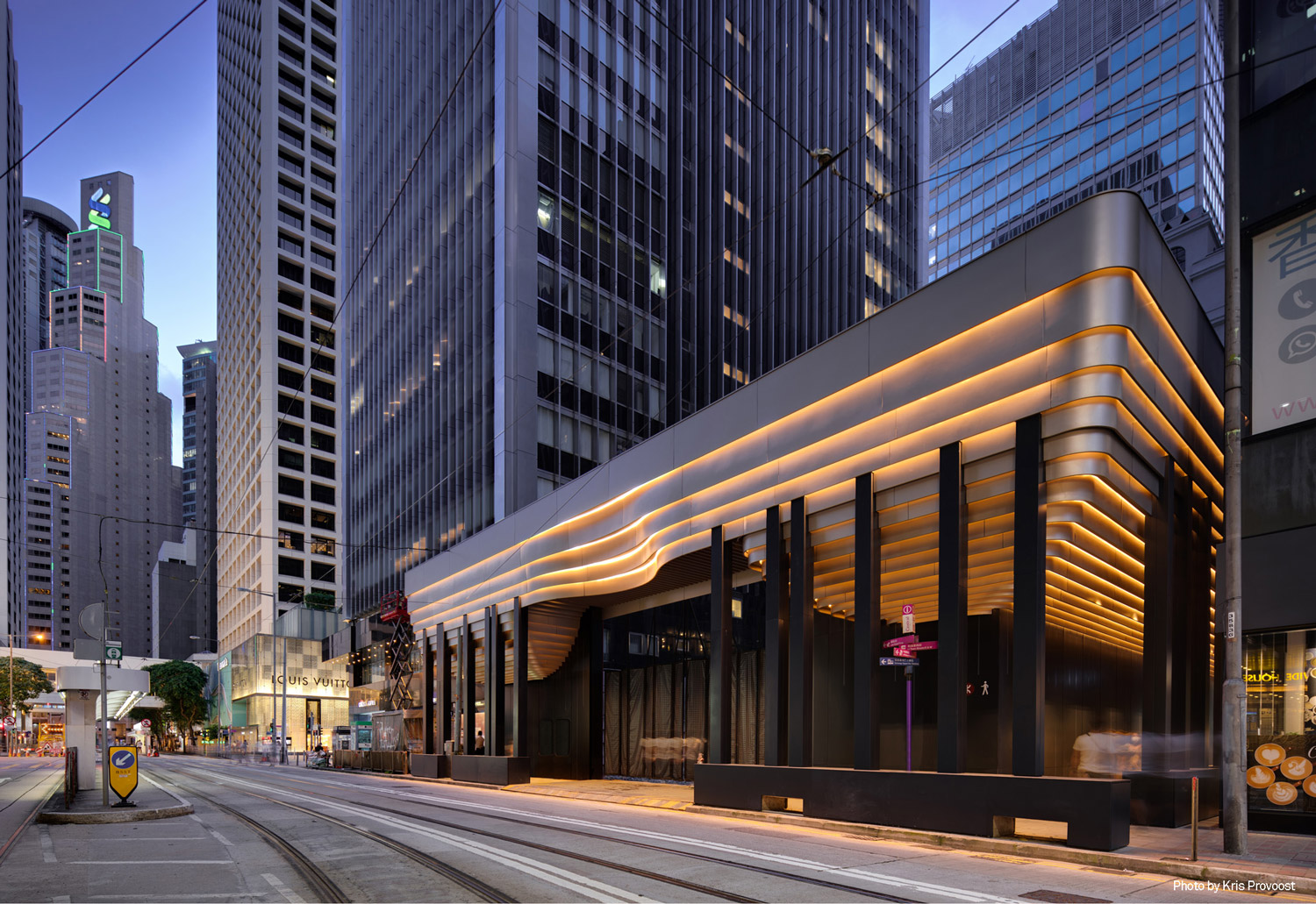 Aedas’ Takshing House Redevelopment construction hoarding in Hong Kong is shown as a sculptural, illuminated streetscape installation with smooth curved aluminium panels, warm linear lighting bands, and rounded corners. The image highlights an architecturally expressive hoarding that engages the CBD context, using flowing forms and a recessed concave entrance gesture to signal the forthcoming landmark redevelopment while complementing the surrounding urban fabric at Theatre Lane.