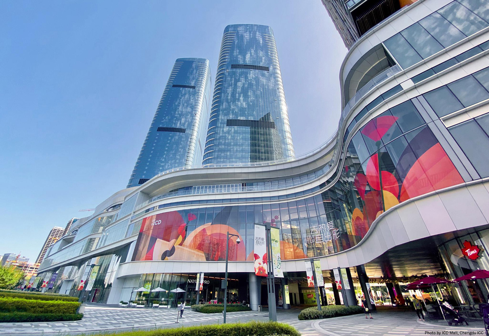 Aedas’ ICD Mall at Chengdu International Commerce Centre is shown as a fluid, multi‑storey retail podium with sweeping, wave‑like façades and expansive glazed shopfronts beneath rising office towers. The image highlights terraced curves inspired by rivers and natural landscapes, integrating outdoor dining decks, vibrant graphic walls, and landscaped edges to create a relaxed, water‑themed urban shopping destination at the gateway of the Jinjiang Business District.