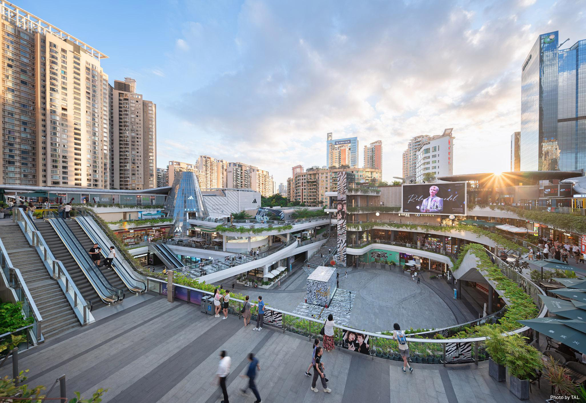 Aedas’ Futian COCO Park in Shenzhen is shown as a revitalised open‑air retail and leisure complex with terraced decks, cascading escalators, and a central sunken plaza surrounded by greenery. The image highlights a park‑like urban environment where fluid circulation, rooftop gardens, and layered public spaces integrate shopping, dining, and social activity within a vibrant city setting.