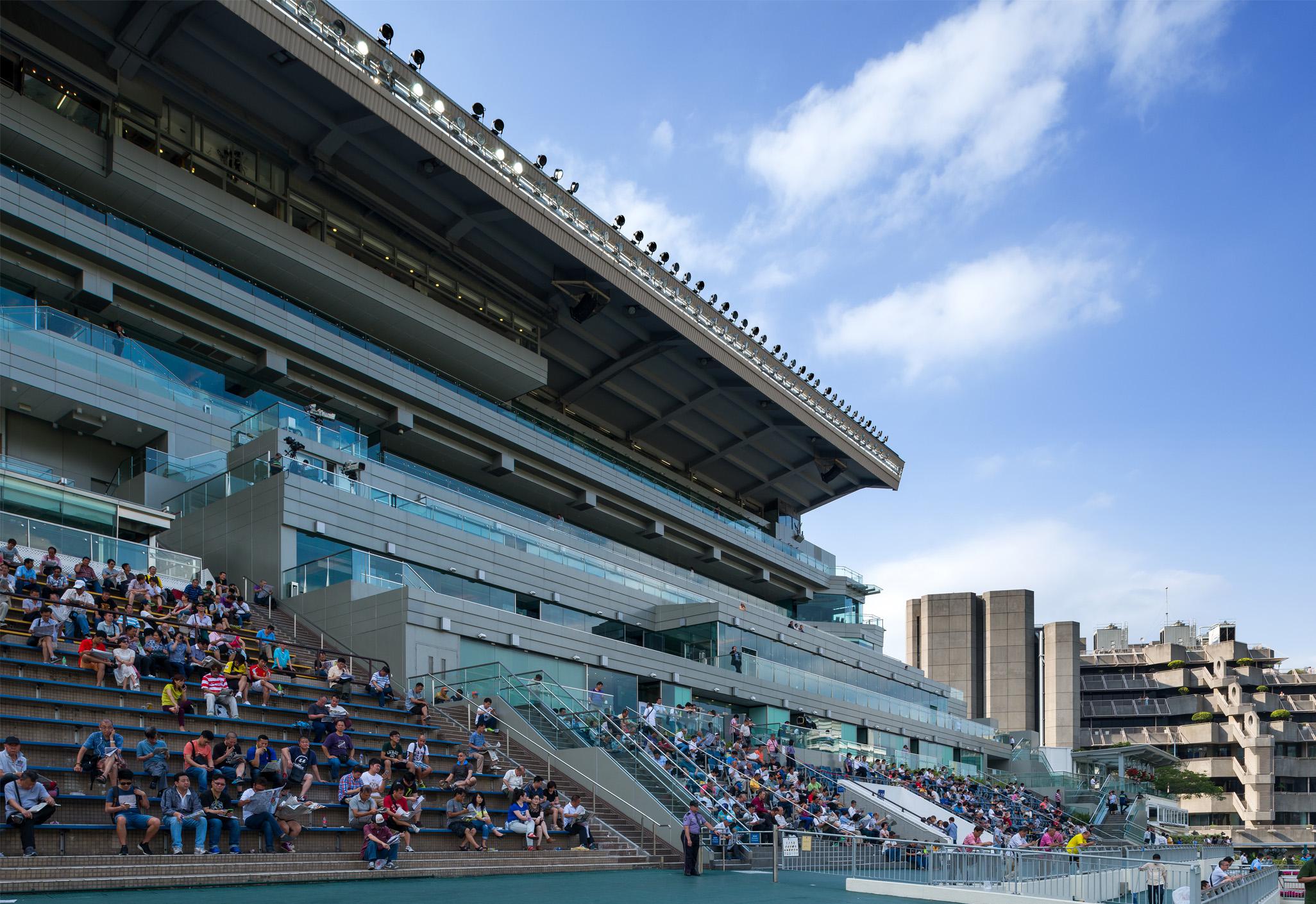 Aedas’ Hong Kong Jockey Club Happy Valley Racecourse renovation is shown as a modernised grandstand with stepped spectator seating, extensive glass balustrades, and a deep cantilevered roof canopy. The image highlights upgraded viewing terraces, contemporary steel and glass architecture, and enhanced public amenities that integrate seamlessly with the historic racecourse, providing panoramic track views and an improved spectator experience within a dense urban setting.