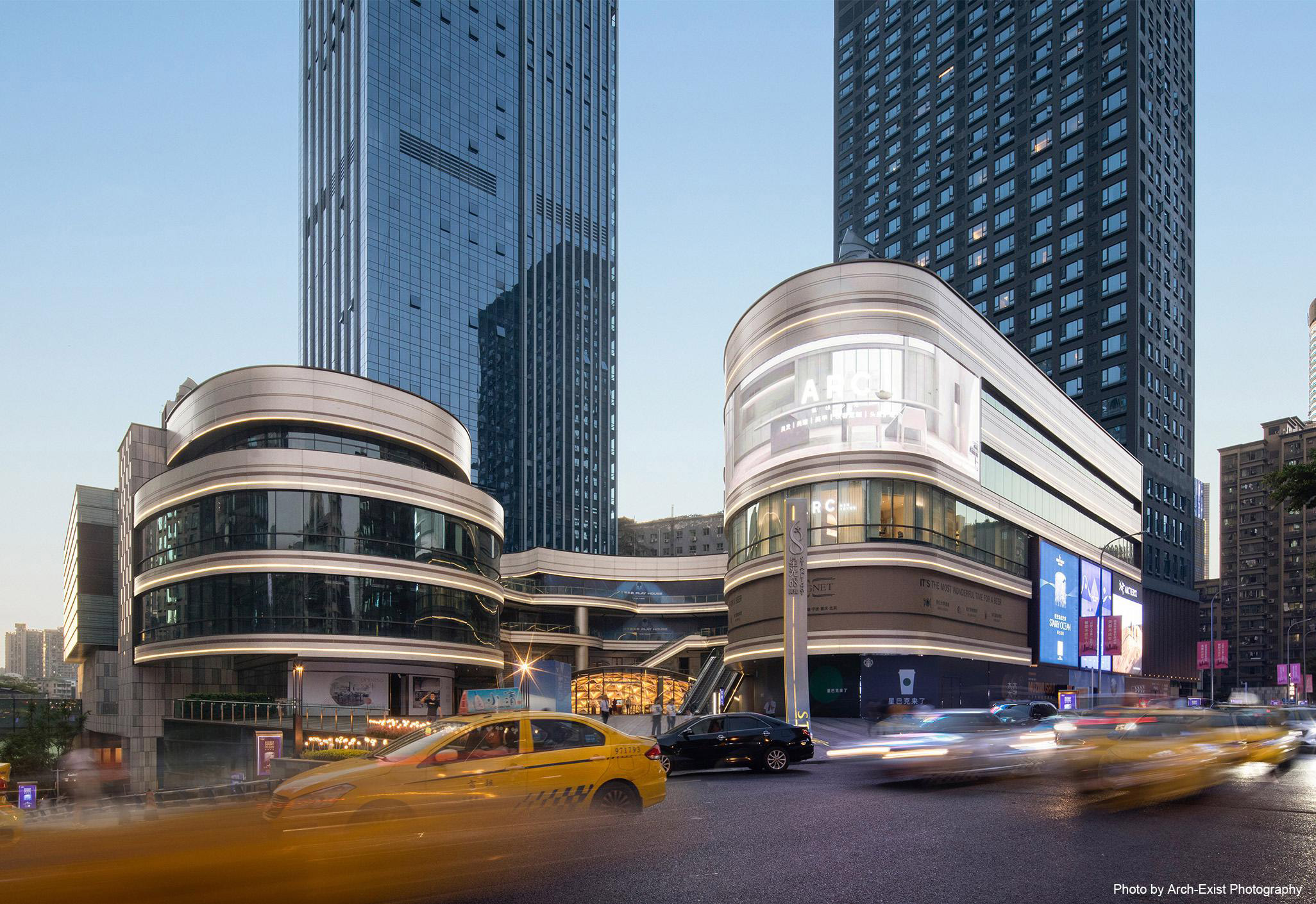 Aedas’ Chongqing Guanyinqiao Sincere Centre Phase II is shown as a dramatic mixed‑use urban complex with a sweeping, crystal‑like retail podium bridging between two high‑rise towers. The image highlights curved glass façades layered with stone and metal fins, illuminated shopfronts, and a dynamic street‑level interface that resolves significant topographical change while creating an immersive, landmark retail and commercial destination within Chongqing’s dense business district.