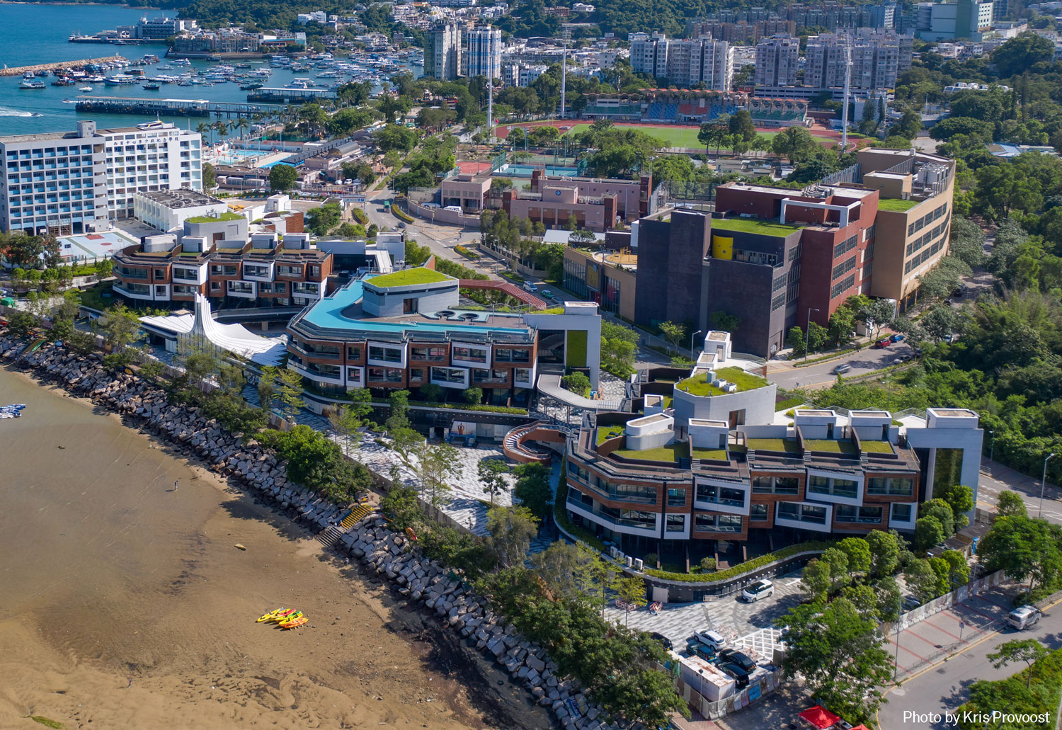 Aedas’ Sai Kung WM Hotel with low‑rise zig‑zag blocks, green roofs and seafront plazas on the Sai Kung shoreline. Aerial view of the Aedas‑designed Sai Kung WM Hotel in Hong Kong, showing three low‑rise hotel blocks arranged in a zig‑zag formation along the coastline. The development features green roof terraces, stepped seafront plazas, curved circulation paths and a waterfront edge integrated with the rocky shoreline and calm bay waters of the Sai Kung Peninsula.