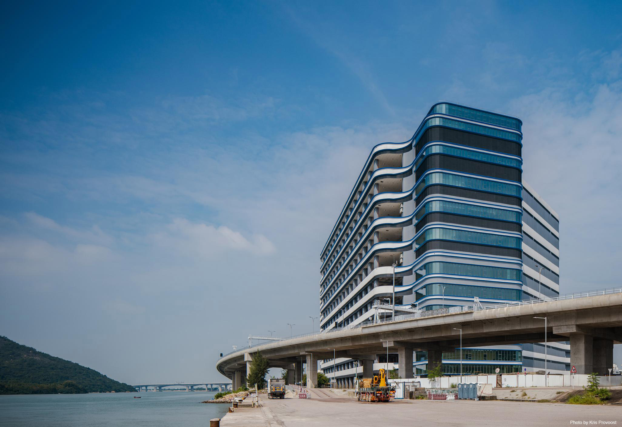 Aedas’s CaiNiao Smart Gateway in Hong Kong is shown as a high‑rise logistics and office building with layered curved façades, continuous horizontal glazing bands, and a podium interface adjacent to transport infrastructure. The image highlights large‑scale logistics architecture integrated with waterfront conditions and elevated road systems.