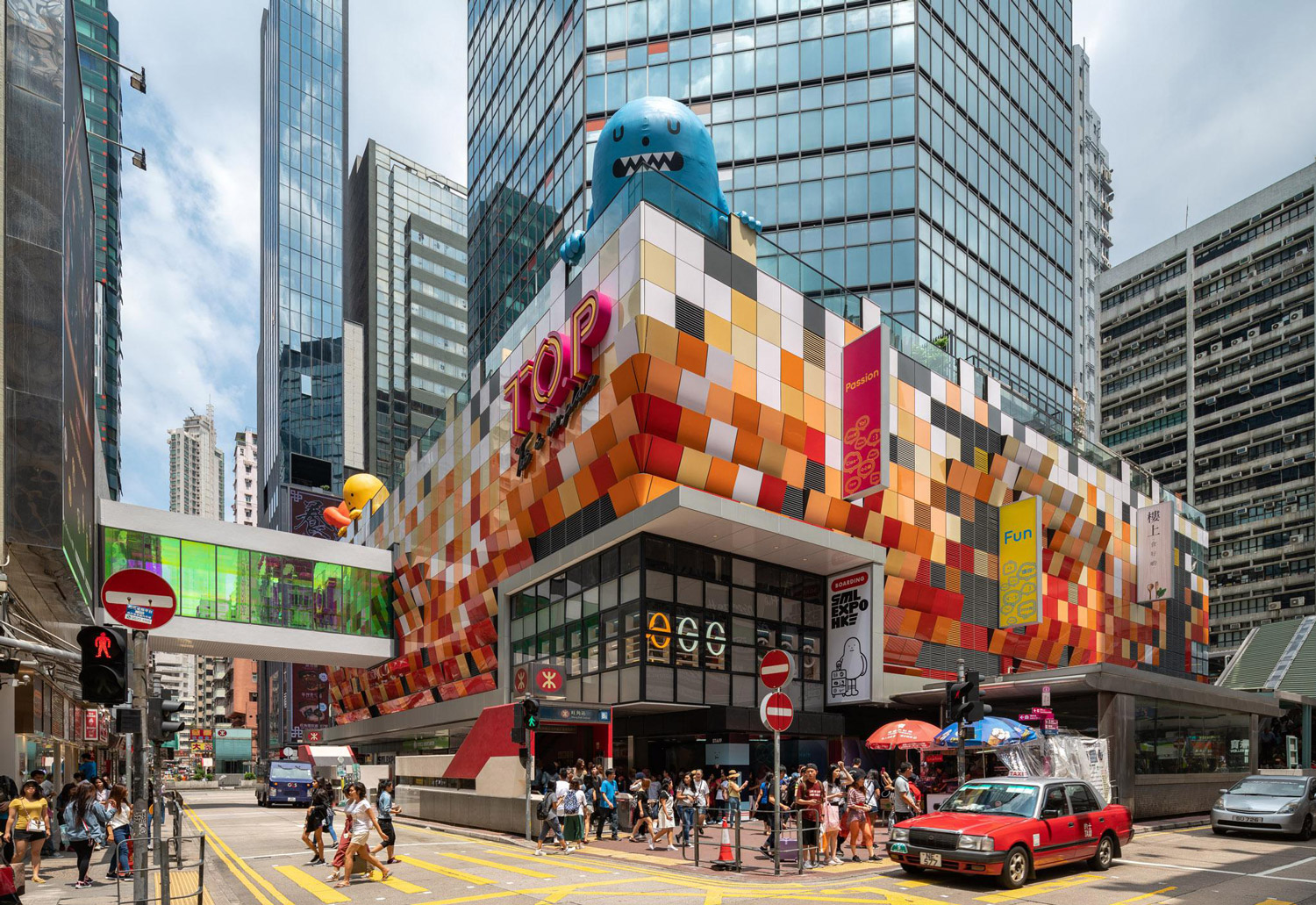 Aedas’ T.O.P – This Is Our Place in Hong Kong is shown as a vibrant urban retail and entertainment hub with a pixelated, multi‑colour podium façade wrapping the street corner. The image highlights a playful architectural expression with bold graphic panels, large glazed openings, overhead pedestrian links, and animated public activity, transforming a former government building into a youthful, contemporary social destination anchored around an open atrium and performance‑oriented public spaces.