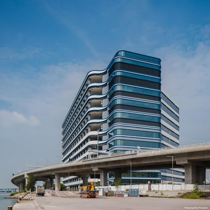 Aedas’s CaiNiao Smart Gateway in Hong Kong is shown as a high‑rise logistics and office building with layered curved façades, continuous horizontal glazing bands, and a podium interface adjacent to transport infrastructure. The image highlights large‑scale logistics architecture integrated with waterfront conditions and elevated road systems.