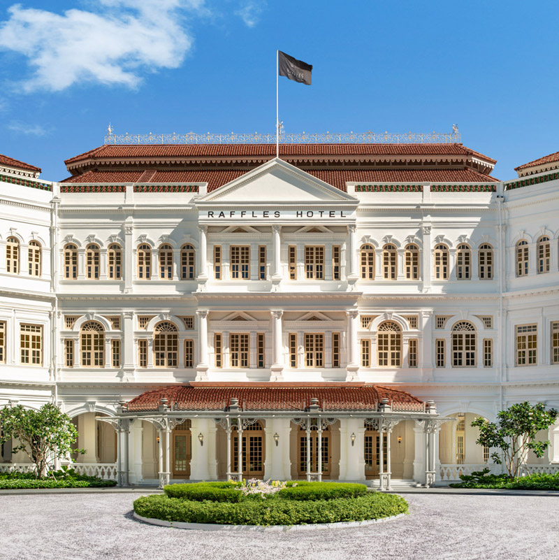 Aedas’ restoration of Raffles Hotel Singapore is shown as the iconic neoclassical façade with white colonnades, arched windows, and red‑tiled roofs set within lush tropical landscaping. The image highlights the carefully preserved heritage architecture, symmetrical proportions, and refined detailing that reflect the hotel’s timeless elegance, gracefully reintegrated with modern upgrades to sustain its legacy as a civic and hospitality landmark.