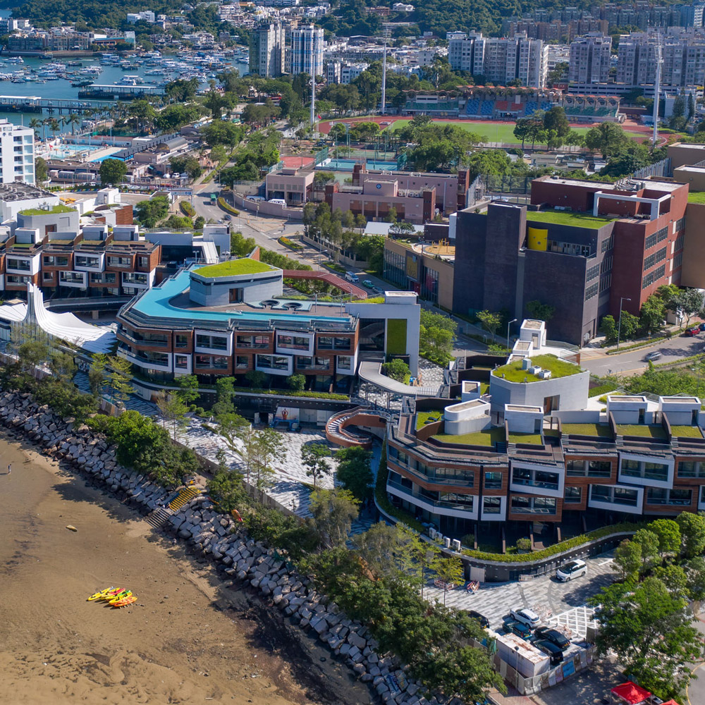 Aedas’ Sai Kung WM Hotel with low‑rise zig‑zag blocks, green roofs and seafront plazas on the Sai Kung shoreline. Aerial view of the Aedas‑designed Sai Kung WM Hotel in Hong Kong, showing three low‑rise hotel blocks arranged in a zig‑zag formation along the coastline. The development features green roof terraces, stepped seafront plazas, curved circulation paths and a waterfront edge integrated with the rocky shoreline and calm bay waters of the Sai Kung Peninsula.