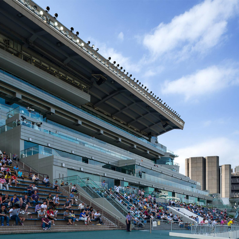 Aedas’ Hong Kong Jockey Club Happy Valley Racecourse renovation is shown as a modernised grandstand with stepped spectator seating, extensive glass balustrades, and a deep cantilevered roof canopy. The image highlights upgraded viewing terraces, contemporary steel and glass architecture, and enhanced public amenities that integrate seamlessly with the historic racecourse, providing panoramic track views and an improved spectator experience within a dense urban setting.