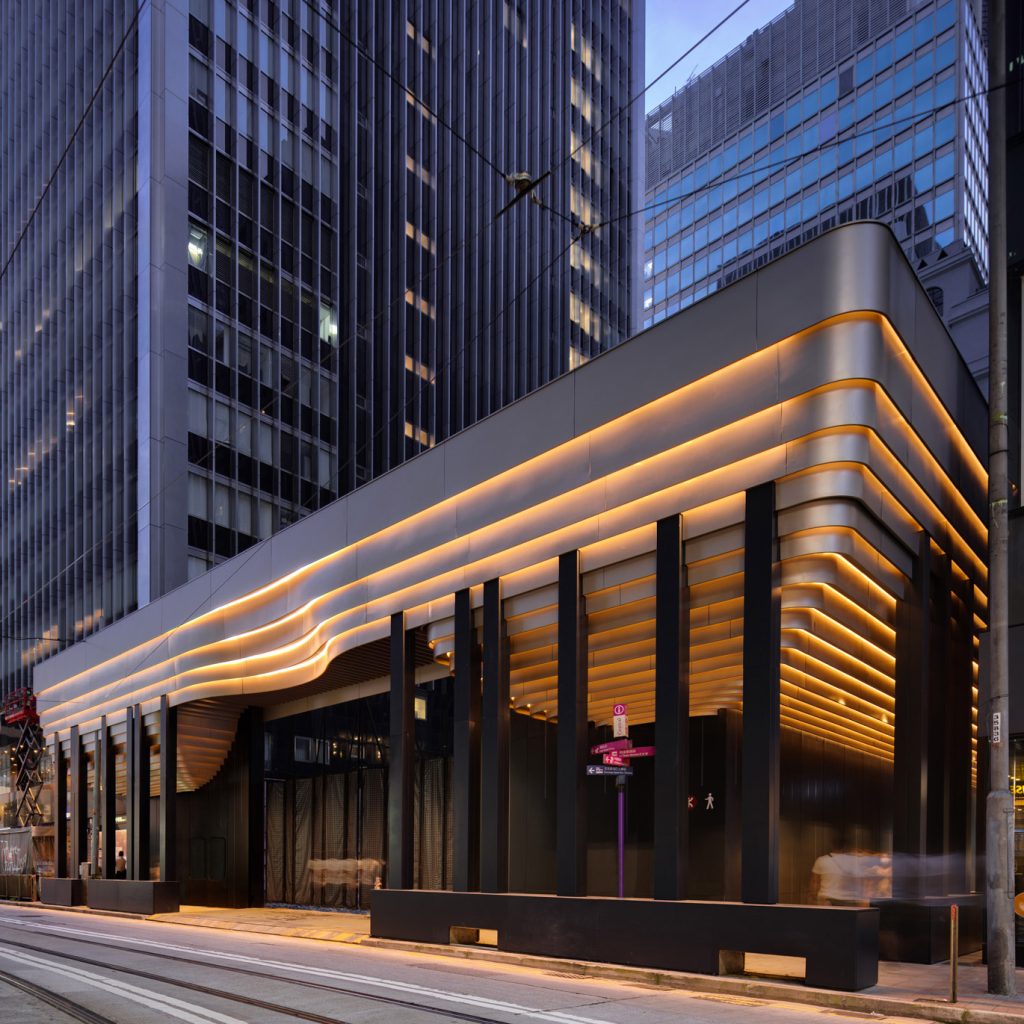 Aedas’ Takshing House Redevelopment construction hoarding in Hong Kong is shown as a sculptural, illuminated streetscape installation with smooth curved aluminium panels, warm linear lighting bands, and rounded corners. The image highlights an architecturally expressive hoarding that engages the CBD context, using flowing forms and a recessed concave entrance gesture to signal the forthcoming landmark redevelopment while complementing the surrounding urban fabric at Theatre Lane.