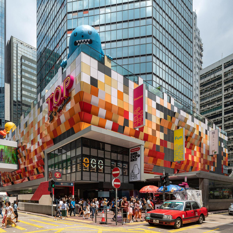 Aedas’ T.O.P – This Is Our Place in Hong Kong is shown as a vibrant urban retail and entertainment hub with a pixelated, multi‑colour podium façade wrapping the street corner. The image highlights a playful architectural expression with bold graphic panels, large glazed openings, overhead pedestrian links, and animated public activity, transforming a former government building into a youthful, contemporary social destination anchored around an open atrium and performance‑oriented public spaces.