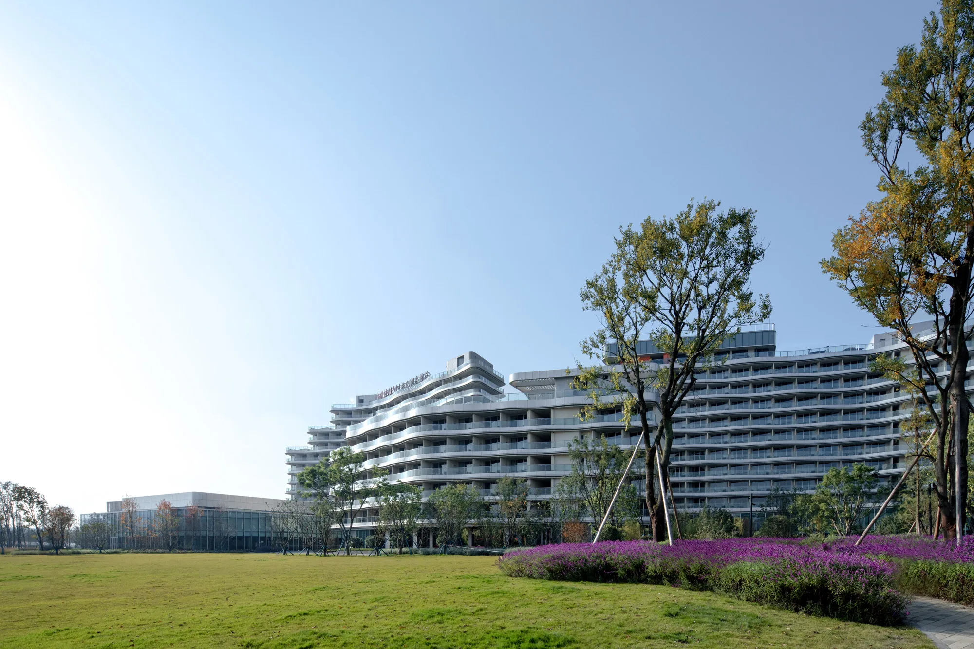 Aedas’ Mumian Chengdu Dong’an Lake is shown as a low‑rise, horizontally layered resort building with cascading terraces extending toward an expansive lakeside park. The image highlights a jagged, linear architectural form integrated with landscaped lawns and mature trees, creating a tranquil leisure destination that connects hotel, public plaza, and ecological surroundings while framing views of Dong’an Lake and the distant Longquan Mountains.