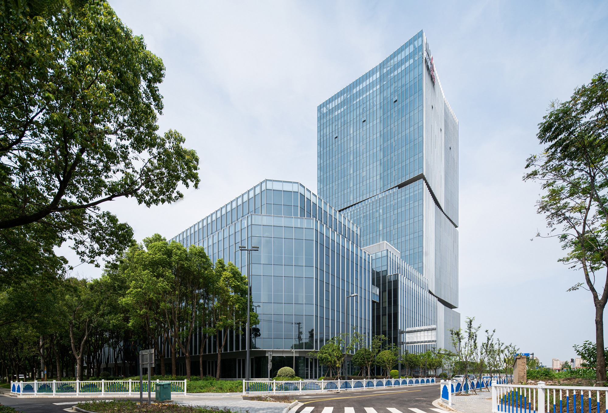 Aedas’ Shanghai Jiading Financial Center is shown as a contemporary office campus with a slender Grade‑A tower and lower‑rise office blocks set within dense greenery. The image highlights a crisp glass façade with articulated vertical fins, inward‑tapered sides, and outward‑extended corners, forming a light, elegant skyline that integrates seamlessly with the surrounding ecological landscape of Jiading New City.
