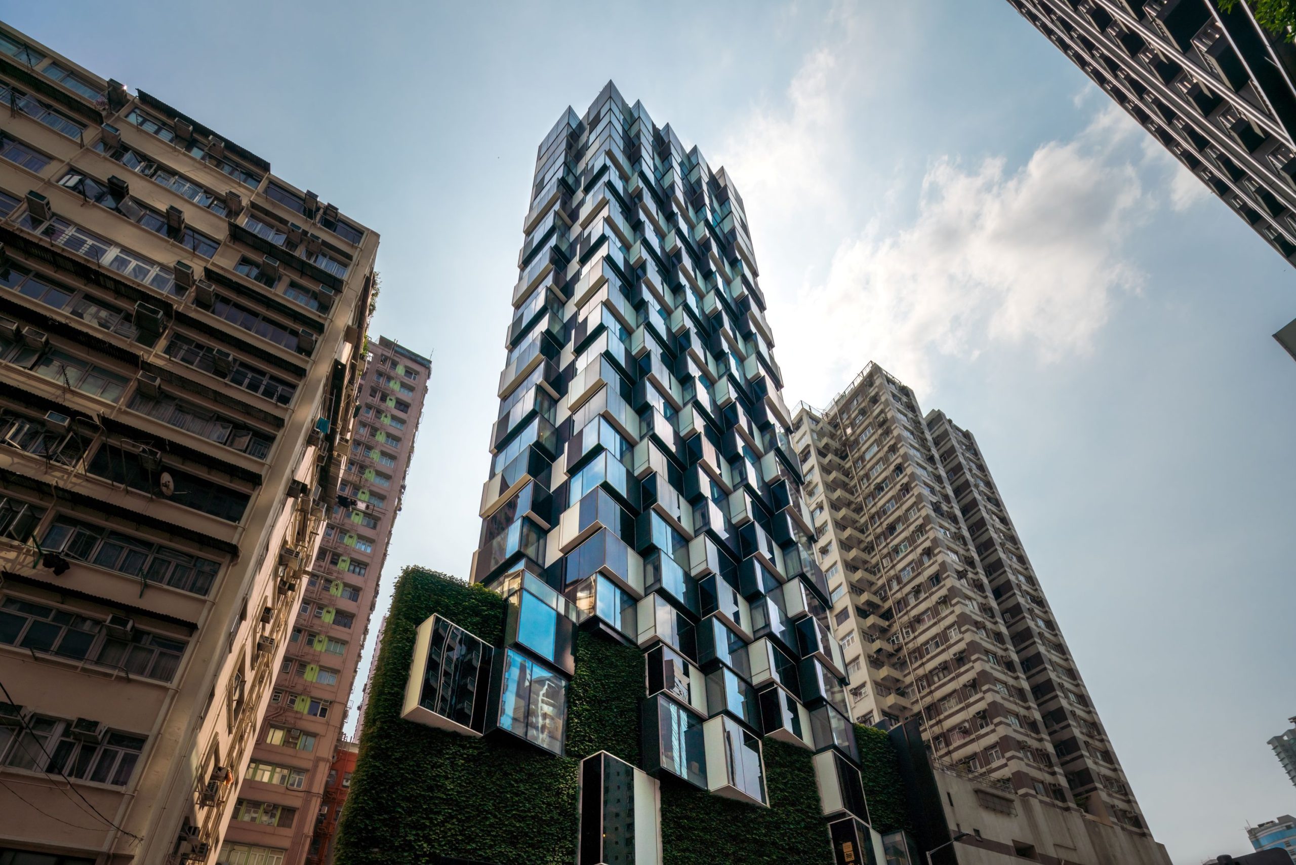 Aedas’s The Beacon in Hong Kong is shown as a slender residential high‑rise with a faceted glass façade, projecting box‑like window bays, and a planted podium base. The exterior highlights sculptural façade articulation, vertical residential stacking, and integration within a dense urban infill condition typical of Hong Kong.