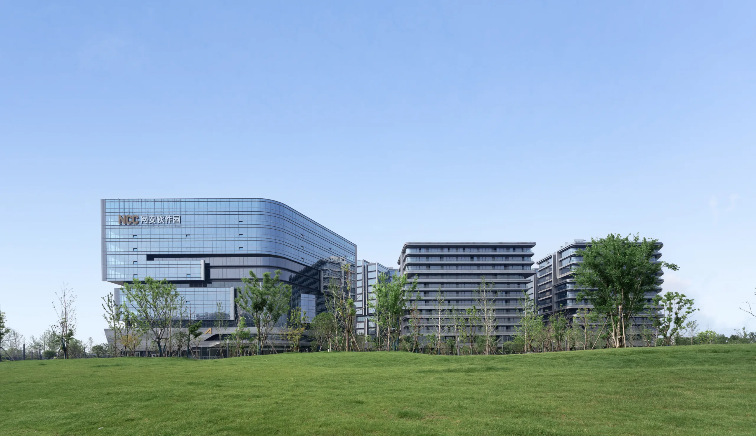 Aedas’s Wuhan National Cybersecurity Center Core Phase 2 is shown as a low‑ to mid‑rise campus development with horizontally layered building volumes, curved glazed façades, and dispersed blocks set within a landscaped green field. The image highlights institutional campus architecture integrating controlled massing, transparent envelopes, and park‑like open space in Wuhan.