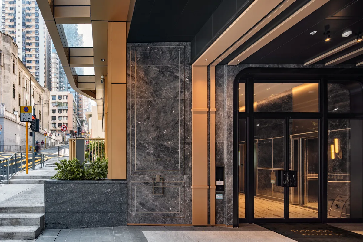 Aedas’ The Nova Residential Development in Hong Kong is shown as a refined residential entrance with stone‑clad walls, bronze‑toned metal framing, and full‑height glazed lobby doors. The image highlights a contemporary urban façade with recessed lighting, layered materials, and landscaped edges, creating an elegant arrival sequence that balances luxury detailing with a calm, modern residential identity within the dense Sai Ying Pun streetscape.