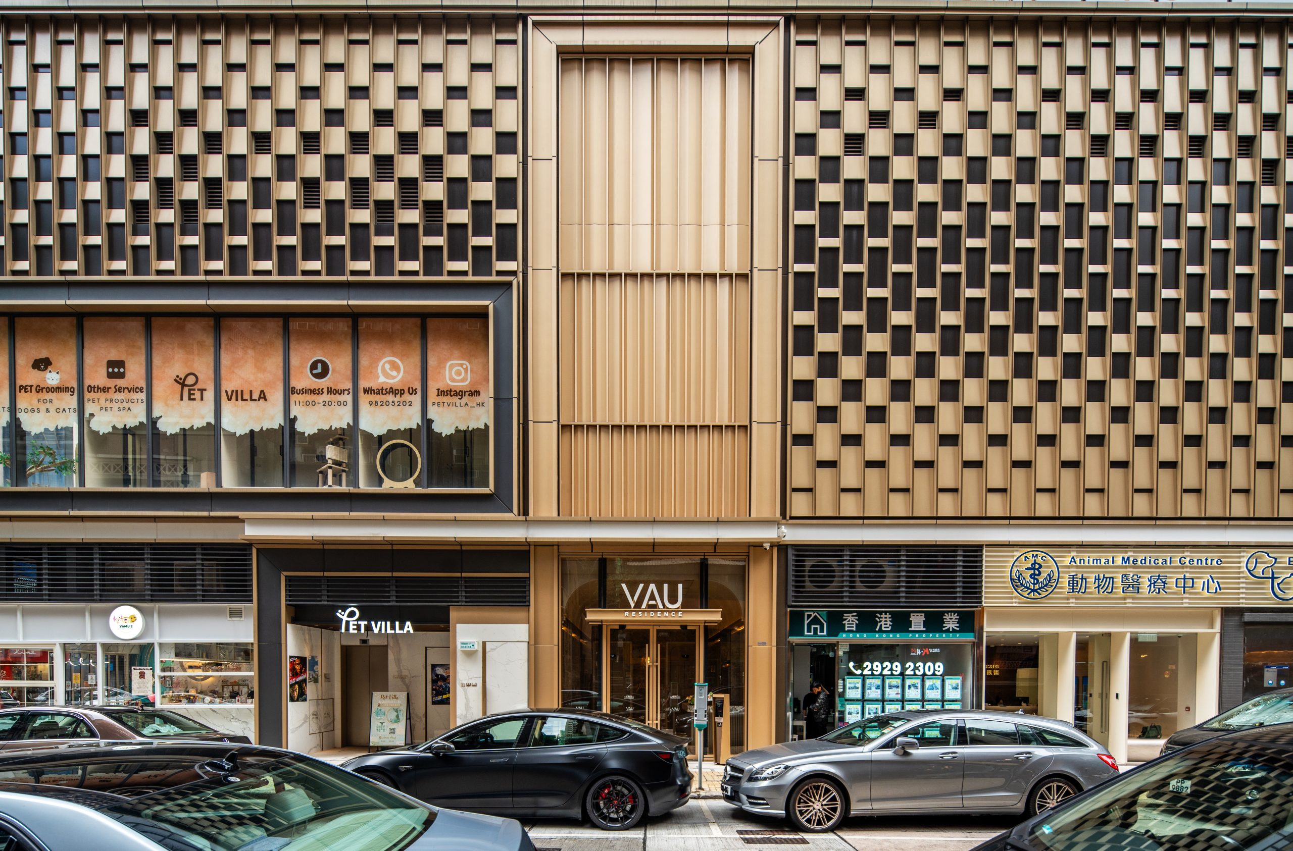Aedas’ VAU Residence in Hong Kong is shown as a contemporary residential tower with a finely articulated podium clad in perforated champagne‑gold aluminium panels and full‑height glazing at street level. The image highlights stacked balconies with glass balustrades, light grey curtain‑wall façades, and metal accents that create rich light‑and‑shadow effects, establishing a refined Southern European‑inspired landmark within the dense Ho Man Tin urban streetscape.
