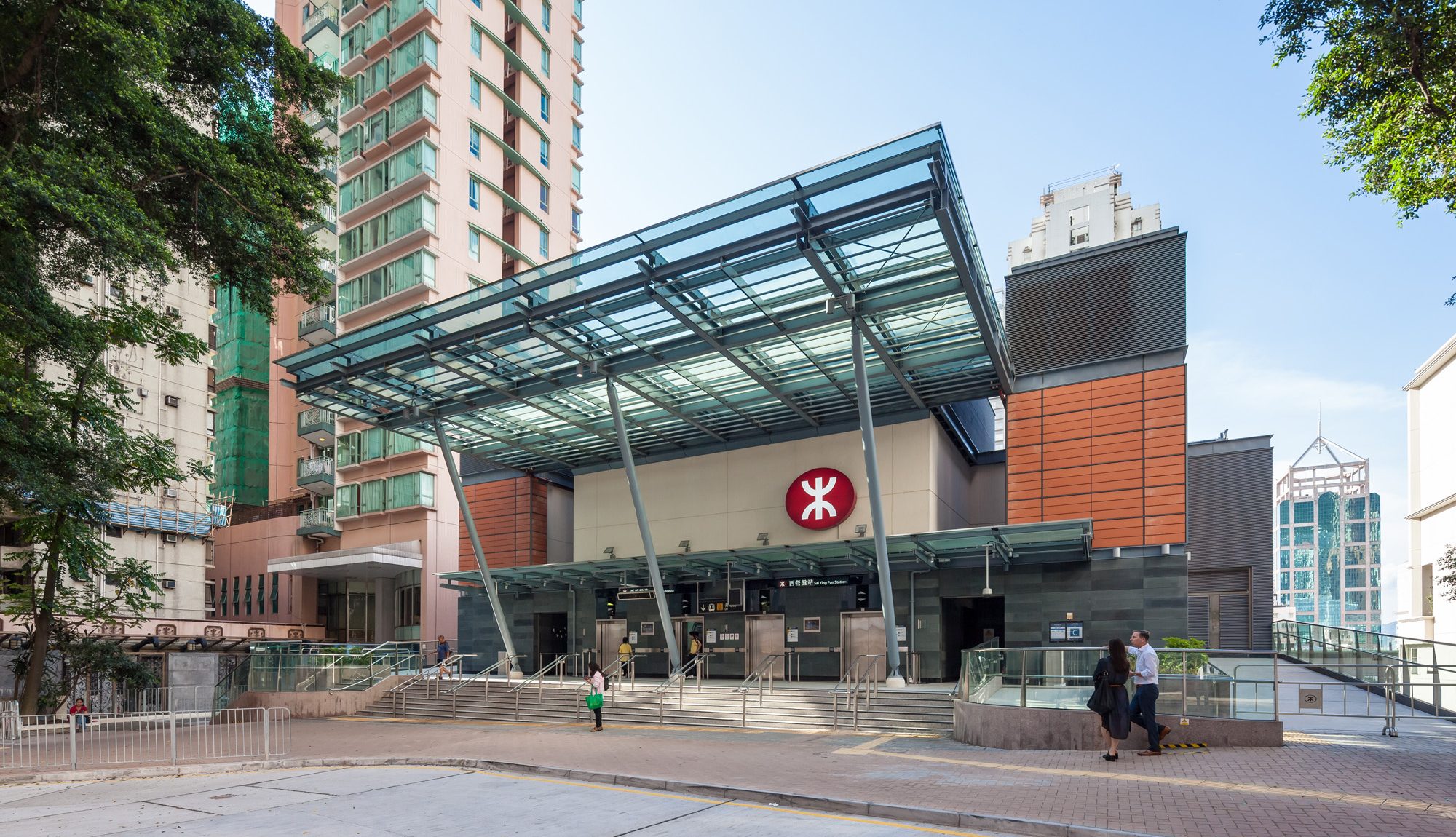 Aedas’s Sai Ying Pun Station on the MTR West Island Line is shown with a transparent glass canopy, exposed steel support frame, stepped entrance plaza, and compact station volume integrated into a dense urban setting. The exterior highlights weather‑protected access, structural clarity, and contemporary metro station architecture in Hong Kong.