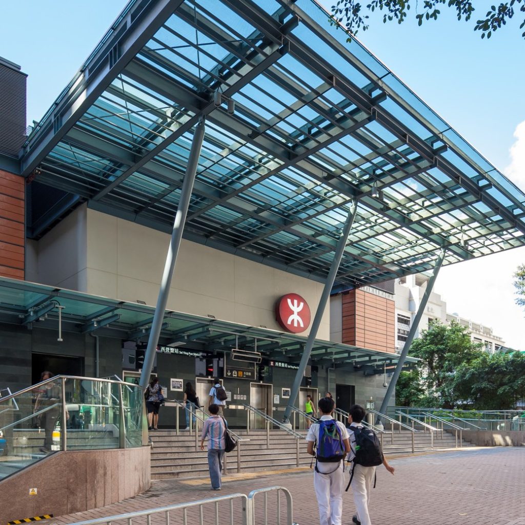Aedas’s Sai Ying Pun Station on the MTR West Island Line is shown with a transparent glass canopy, exposed steel support frame, stepped entrance plaza, and compact station volume integrated into a dense urban setting. The exterior highlights weather‑protected access, structural clarity, and contemporary metro station architecture in Hong Kong.
