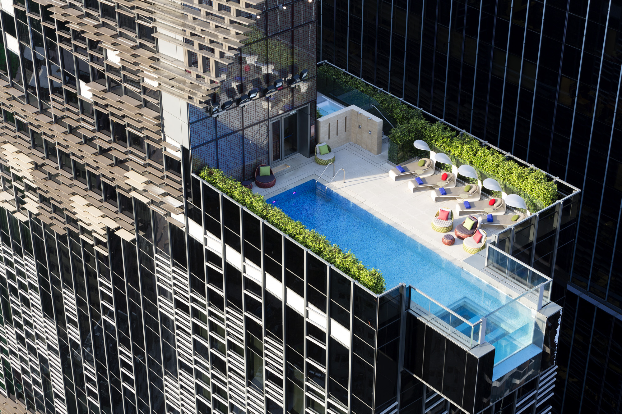 Aedas’s Hotel Indigo in Wan Chai, Hong Kong is shown with a compact rooftop pool terrace enclosed by glass balustrades and planted edges, integrated within a high‑rise hotel tower wrapped in a perforated bronze ecoscreen. The image highlights climate‑responsive façade design, outdoor leisure space, and urban rooftop programming.
