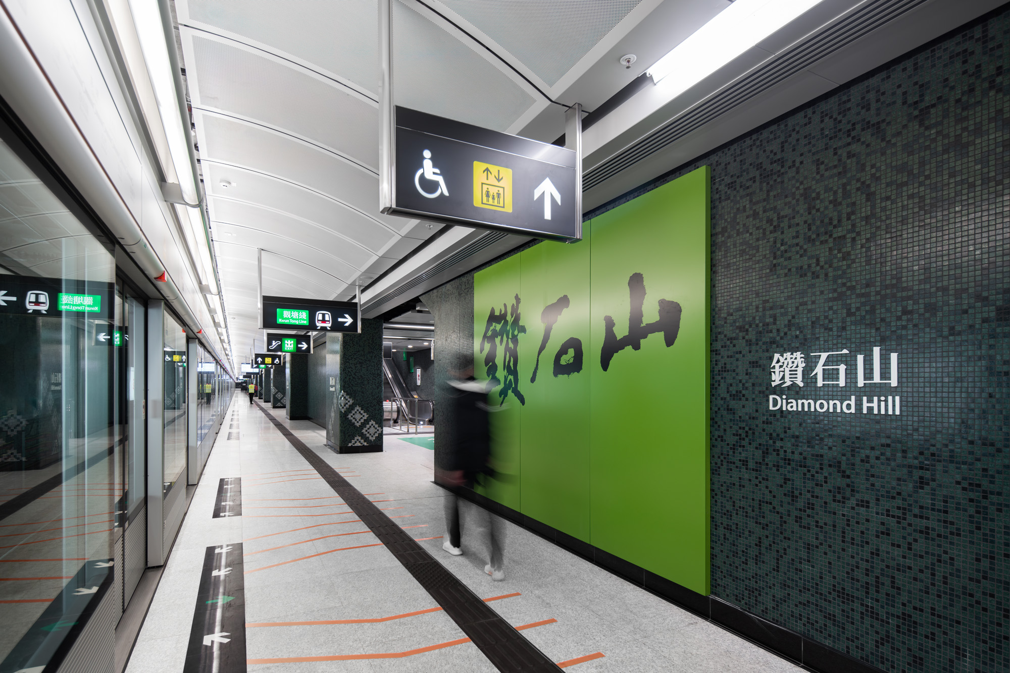 Aedas’s Diamond Hill Station on the MTR Tuen Ma Line is shown with a linear platform environment, curved perforated ceiling panels, integrated lighting, full‑height screen doors, and graphic wall panels marking station identity. The image highlights contemporary metro architecture focused on clarity, robustness, and passenger‑oriented spatial organisation in Hong Kong.