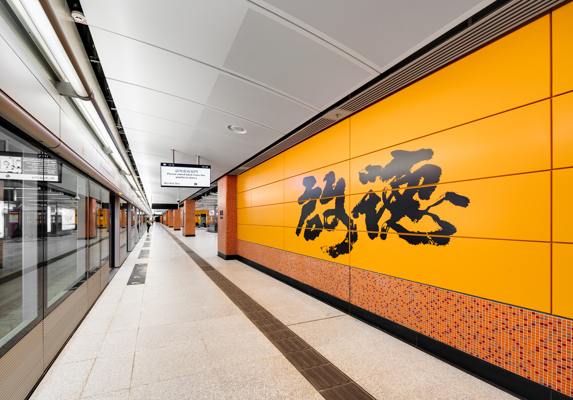Aedas’s Kai Tak Station on the MTR Tuen Ma Line is shown with a linear platform layout, curved perforated ceiling panels, integrated lighting, full‑height platform screen doors, and large‑scale graphic wall finishes. The image highlights contemporary metro architecture focused on clarity, robustness, and passenger‑oriented spatial organisation in Hong Kong.