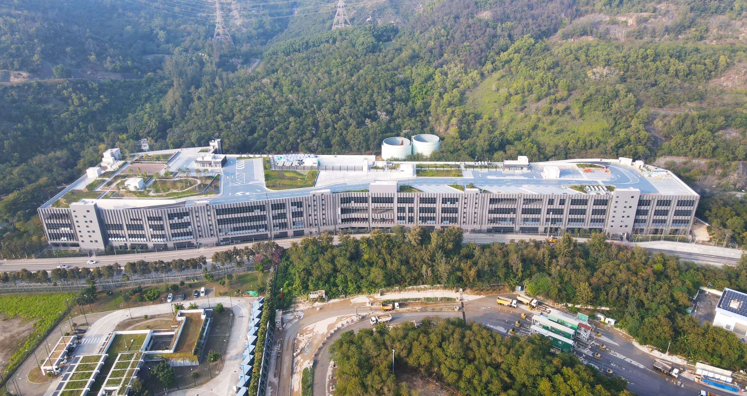 Aedas’s Goodman Westlink Logistics Center in Hong Kong is shown as a large terraced logistics complex with long horizontal floor plates, articulated service façades, expansive rooftop yards, and integrated access roads. The aerial view highlights hillside‑responsive massing and large‑scale industrial architecture embedded within a constrained, green terrain.